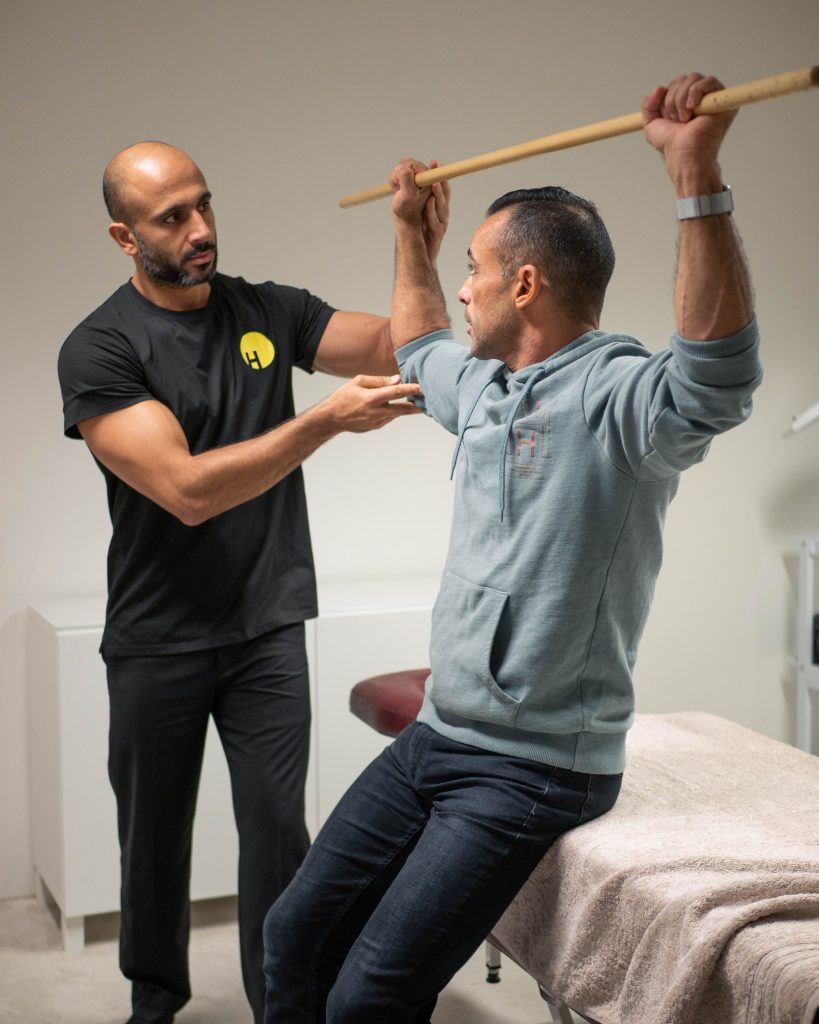 Personal trainer and masseur assisting a sitting person on a bench holding a stretching stick with both hands above the head.